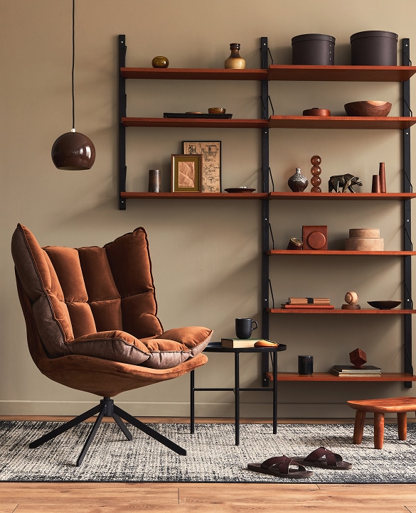 Coin salon cosy avec fauteuil marron, étagères murales bois garnies de décorations, lampe pendante et tapis texturé.