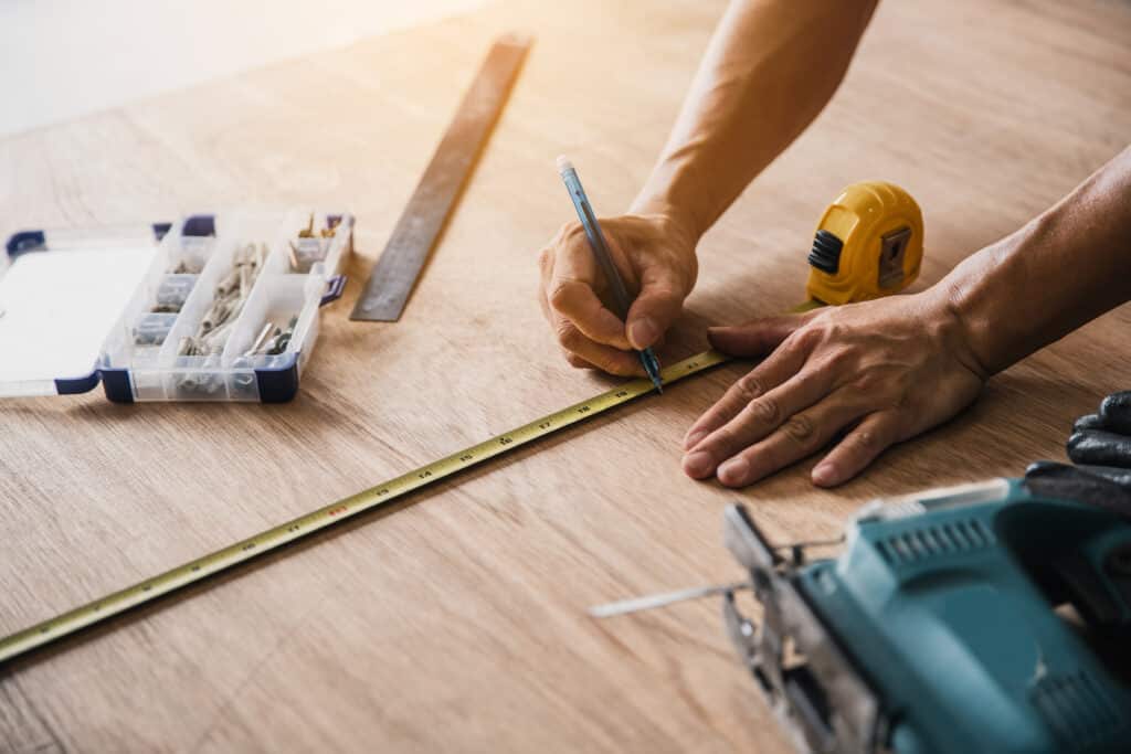 Mains mesurant et marquant une planche de bois avec un mètre ruban et un stylo. Outillage et éléments de fixation visibles.
