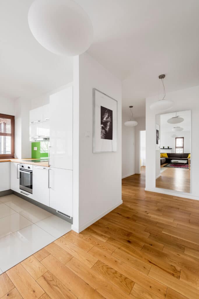 Cuisine moderne blanche avec four intégré, comptoir bois et crédence verte. Parquet clair et miroir mural reflétant le salon lumineux.