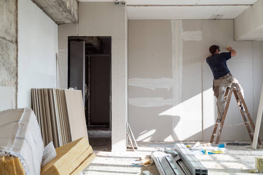 Ouvrier installant des panneaux de placo sur un mur. Chantier de rénovation avec matériaux au sol : isolant, rails. Lumière naturelle.