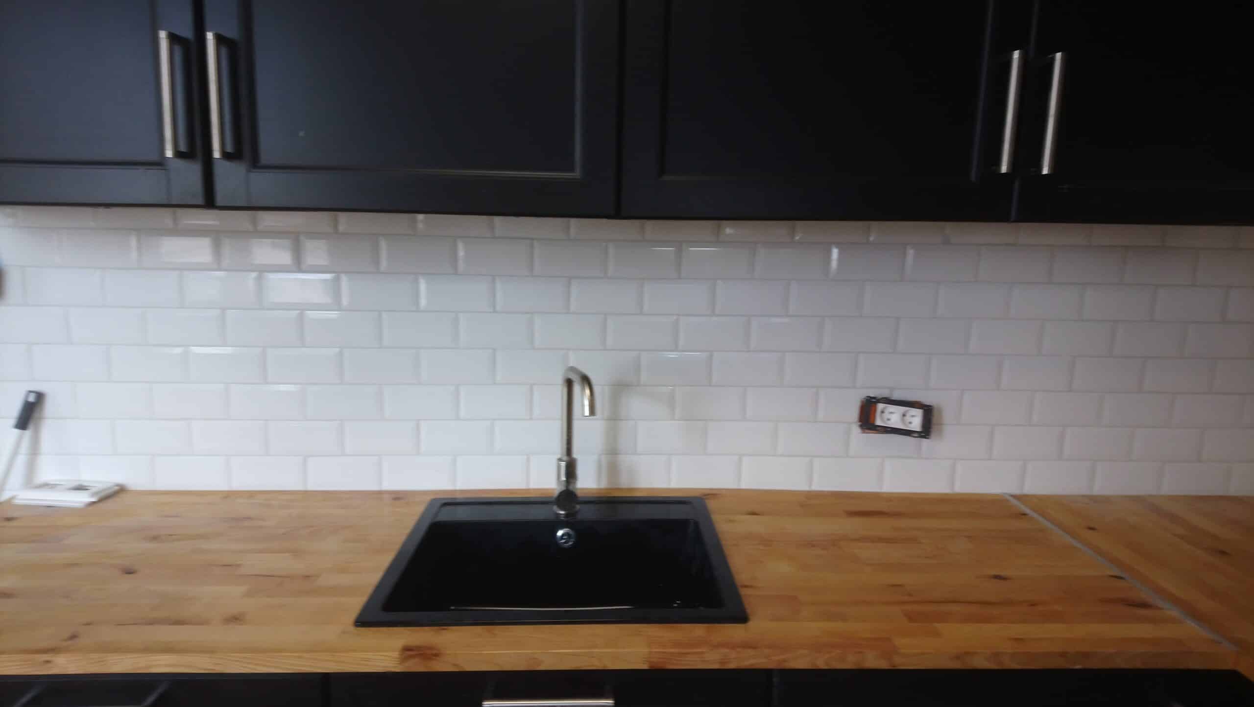 Cuisine avec évier noir encastré dans un plan de travail en bois, crédence métro blanche et meubles hauts noirs.