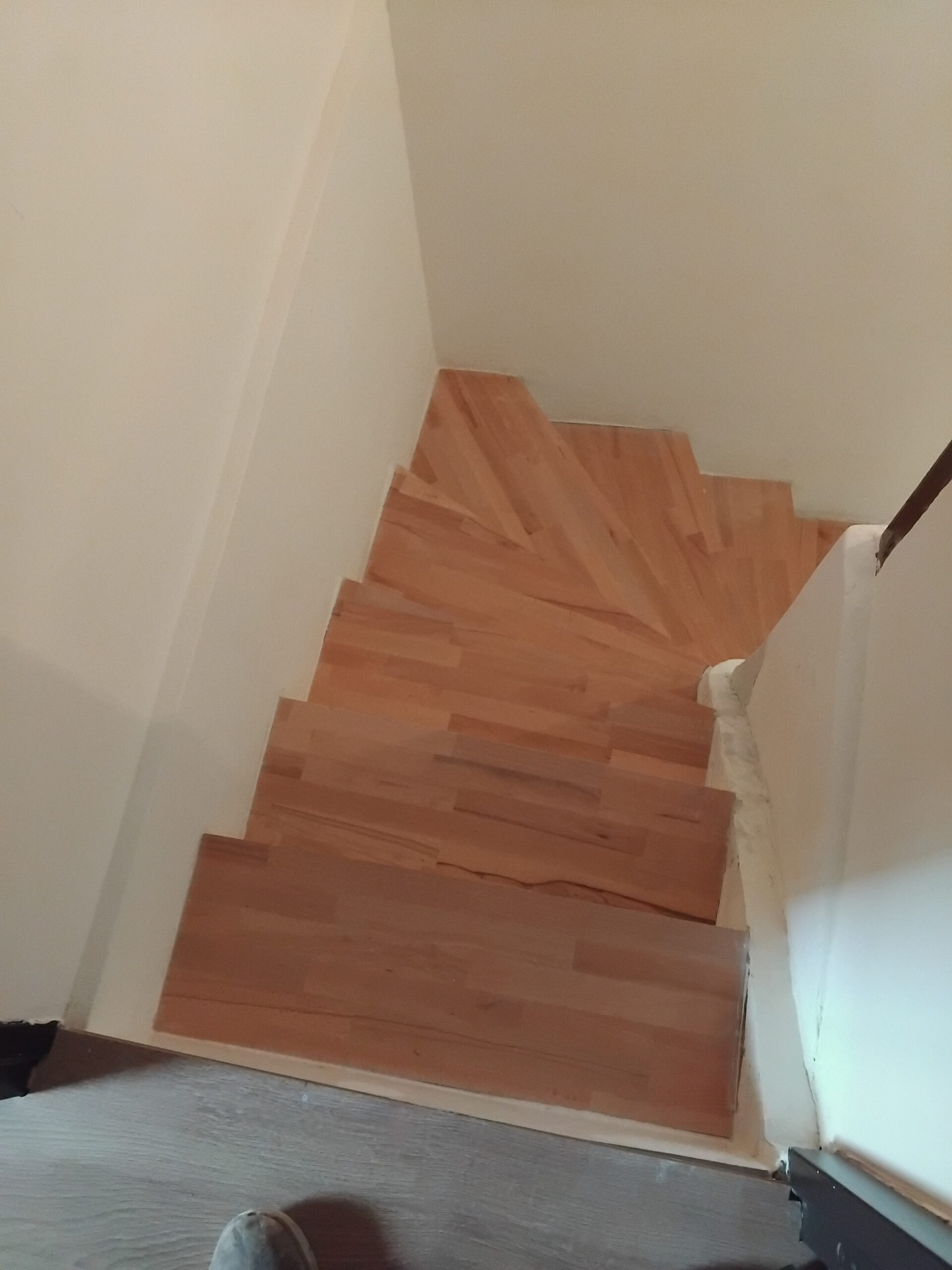 Marches d'escalier en bois clair tournant vue de dessus entre des murs blancs.